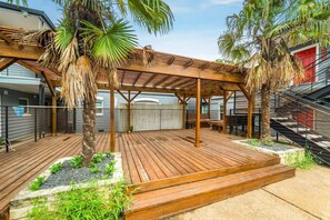 Een houten terras met een pergola, palmbomen en een stenen plantenbak met beplanting.