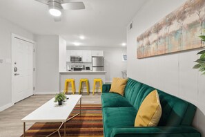 Relax in this stylish living space featuring a green sofa, coffee table, and vibrant rug, alongside a fully equipped kitchen and a unique abstract art piece.