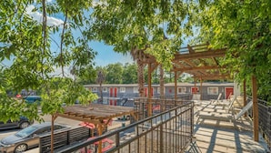 Un espace extérieur aménagé, abrité et doté de tables et de chaises, d’une pergola en bois et d’une vue sur une rangée de bâtiments aux portes rouges.