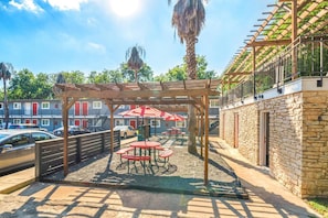 A covered outdoor seating area with tables and umbrellas, adjacent to a stone building with a wooden pergola overhead.