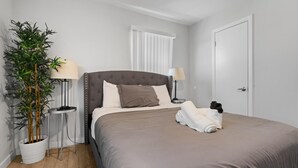 A neatly made bed with a grey headboard, two bedside lamps, a potted plant, and a door. - Modern 2BR Near UT Hyde Park Evonify (Austin)