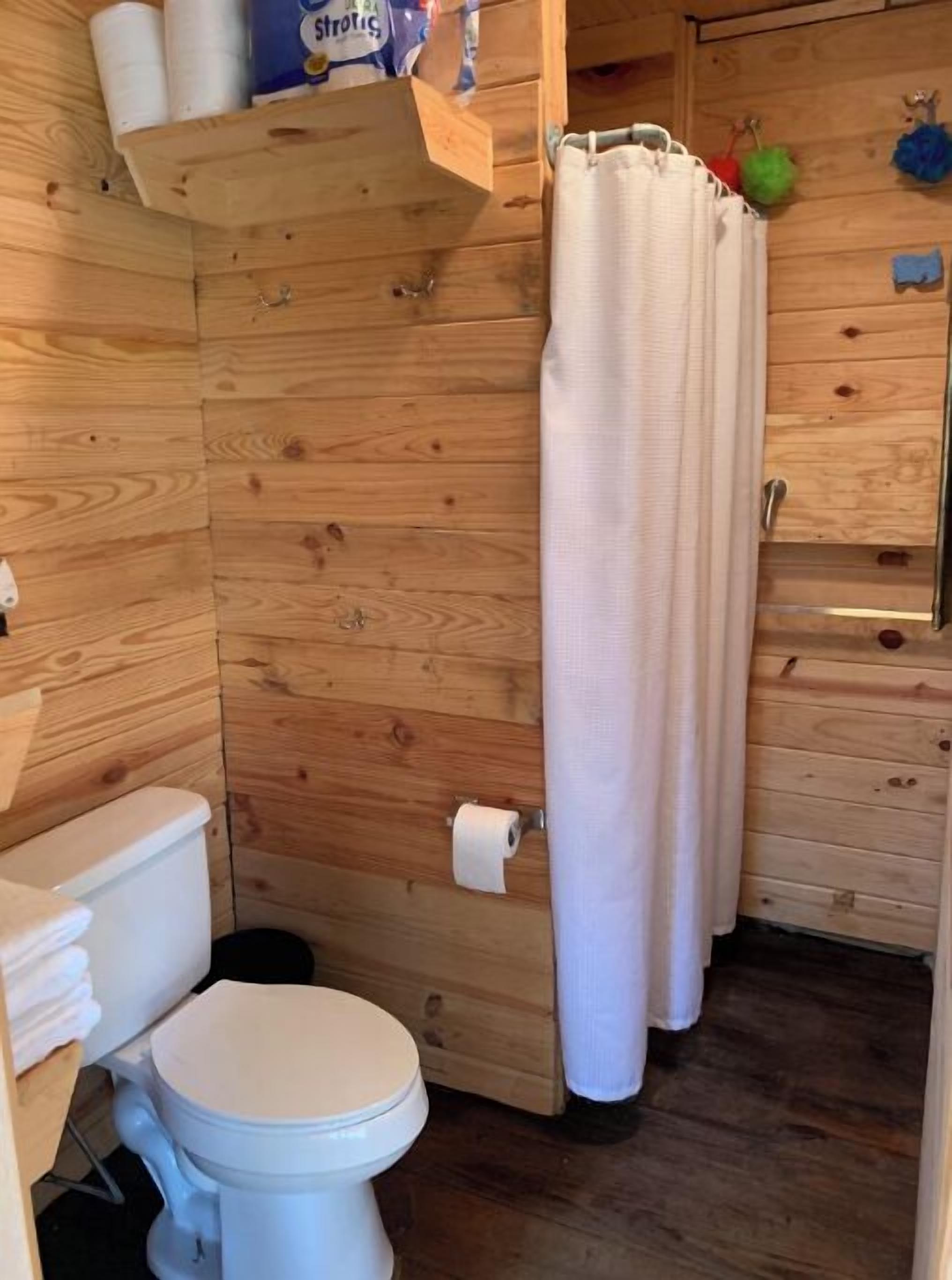 Puisesta mökistä löytyvä kylpyhuone, jossa on wc, hylly wc-paperille ja suihkuverho.