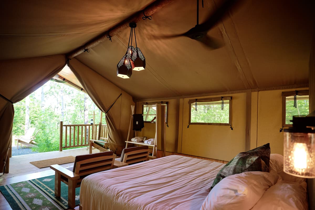 A tented accommodation room with a bed, a small table, a chair, and a window with a view of greenery.