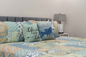 Una cama con varios almohadones decorativos, un cabecero tapizado en gris y una mesita de noche con una lámpara.