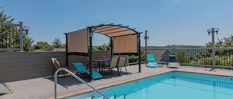 A swimming pool with a wooden deck, lounge chairs, a covered seating area, and a view of the landscape.