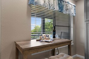 A window with blinds, a wooden table with a bottle, a small house model, and a chair.