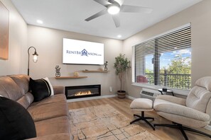 Una sala de estar moderna con chimenea, ventilador de techo, televisor montado y un amplio ventanal con persianas.