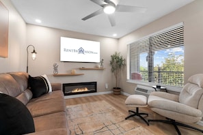 A modern living room with a fireplace, a ceiling fan, a mounted TV, and a large window with blinds.