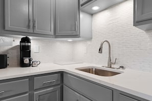 A kitchen with grey cabinets, a white countertop, a stainless steel sink, and a coffee maker.