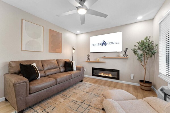 Una sala de estar con un sofá de cuero marrón, chimenea, ventilador de techo y televisor montado mostrando 'RENTBRINSON.COM'.
