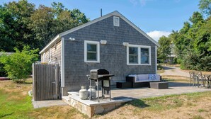 A small, single-story house with a gray exterior, a front porch with a grill, and a bench. - Newly Renovated Cottage on Town Cove! (Eastham)