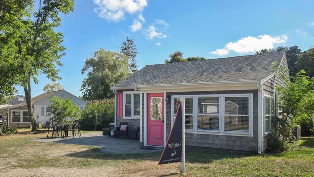A small, single-story building with a red door and windows, surrounded by trees and a grassy area. - Newly Renovated Cottage on Town Cove! (Eastham)