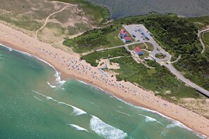 An aerial view of a beach resort with a parking area, a building with a red roof, and a road.