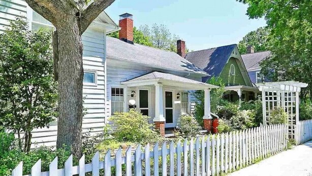A white house with a porch, a picket fence, and a tree in front. - Cozy Historic Oakwood Duplex (Raleigh)