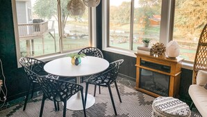 Uma sala de jantar com uma mesa redonda e cadeiras, uma mesinha lateral com livros e uma luminária de sal, e vista para o quintal através de grandes janelas.