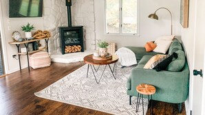 Uma sala de estar acolhedora com um sofá verde, uma mesa de centro de madeira, um tapete estampado, uma parede de pedra, uma lareira e uma janela com vista para árvores.