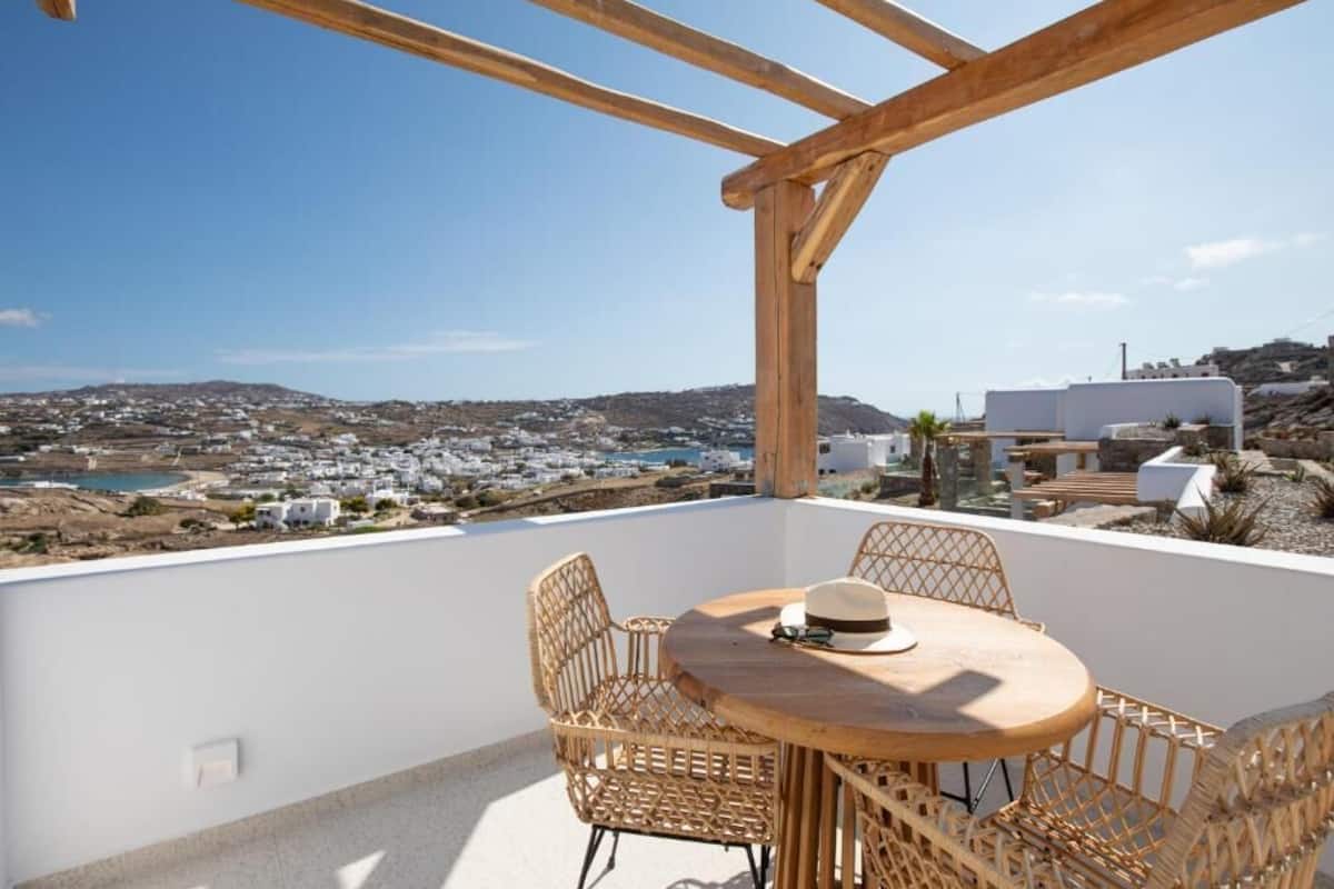 Uma pérgola de madeira com uma mesa redonda e cadeiras de vime, com vista para uma cidade costeira.
