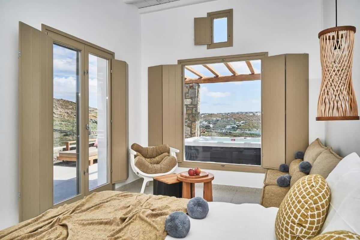 Quarto com uma cama grande, vista para a cidade pela janela, uma cadeira e uma mesinha com frutas.