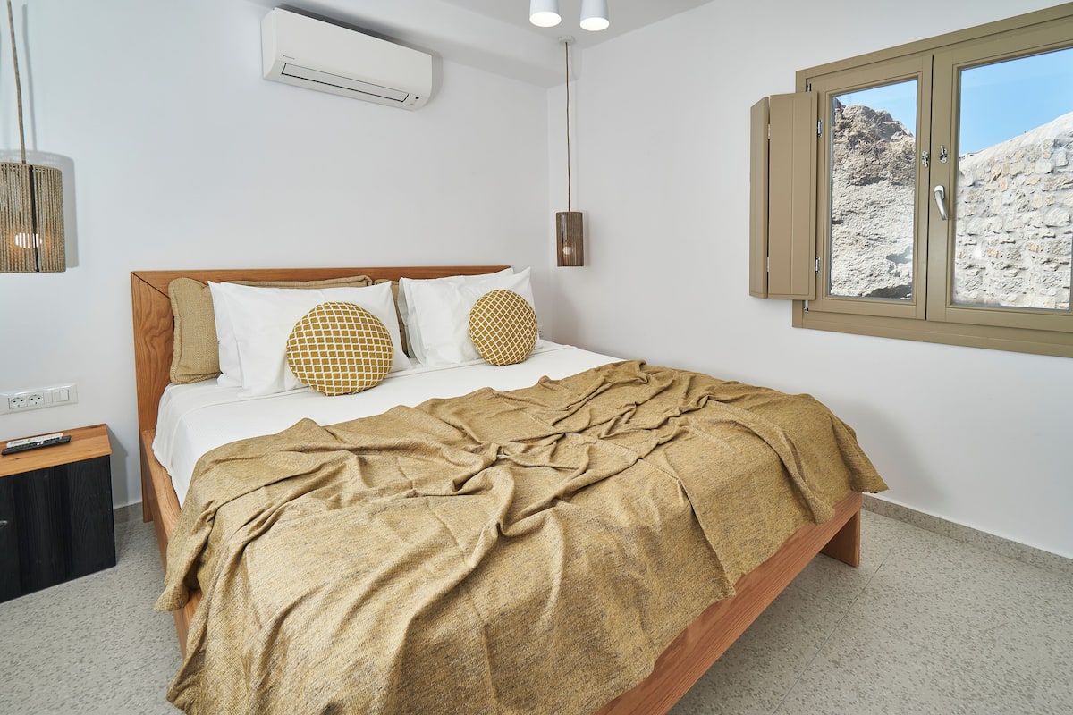 Quarto com cama, janela com vista para uma paisagem rochosa, ar condicionado e mesa de cabeceira com controle remoto.