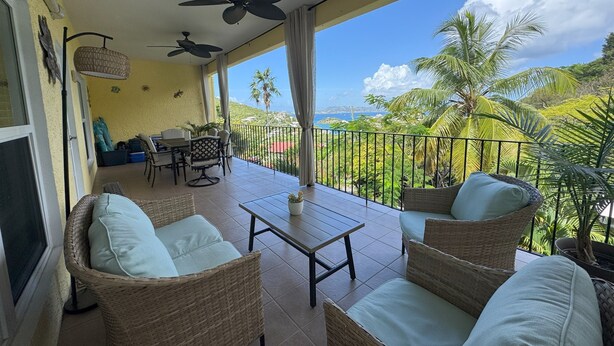 A balcony with wicker furniture, ceiling fans, and a view of palm trees and a distant coastline.