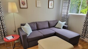 Een moderne woonkamer met een grijze modulaire bank, een kleine rode tafel en een beige voetenbankje. Er hangen drie kunstwerken in lijsten aan de muur en er staat een vloerlamp.