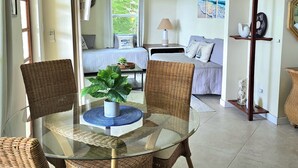Ein Zimmer mit einem Glastisch, umringt von Korbstühlen, einem Bett mit Blick auf Grünflächen, einem Nachttisch mit Lampe und einem an der Wand befestigten Regal mit Dekoration.