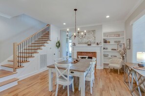A spacious living area with a wooden staircase, a dining table with white chairs, a fireplace, and a bookshelf.