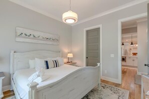 A bedroom with a white bed, a nightstand with a lamp, a painting of birds on the wall, and a view into the kitchen.