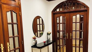 Un couloir avec une porte double en bois, un miroir, une console sur laquelle sont posés un vase et une bougie, et un tapis à motifs.