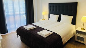 Une chambre d’hôtel avec un grand lit, deux tables de chevet, une fenêtre avec des rideaux légers et une lampe suspendue.