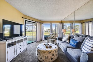 En moderne stue med et fladskærms-tv, en sofa, en stribet lænestol, en rund skammel og store vinduer med udsigt over en golfbane.