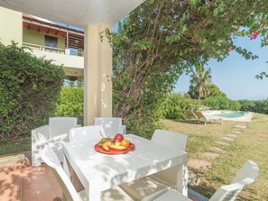 A covered outdoor seating area with a table set with fruit, surrounded by lush greenery and a pool in the background.