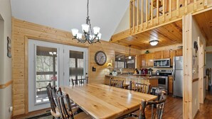 Un interior de madera con una mesa de comedor, sillas, una lámpara de araña, electrodomésticos de cocina y un reloj en la pared.