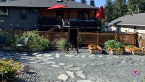 A two-story wooden house with a balcony, a red umbrella, and a gravel patio with stepping stones and flower beds.