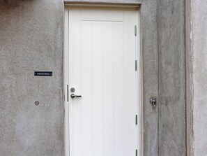 Una puerta blanca con picaporte metálico y cerradura, un pequeño letrero encima, y un muro de concreto.