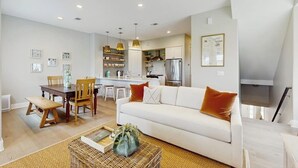 A modern living room with a white sofa, wooden dining table, and a kitchen in the background. - Luxury townhome with games, high-speed WiFi, pool access, AC & beach service (Port Aransas)