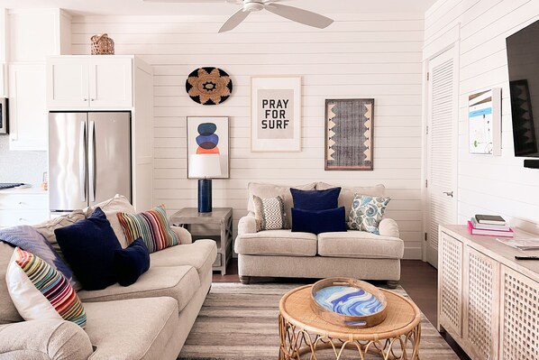 A modern kitchen with white cabinetry, a stainless steel refrigerator, and a dining area with a wicker table and a blue and white patterned bowl. - Townhouse + loft with private balcony, heated pool, gym, beach access & W/D (Port Aransas)