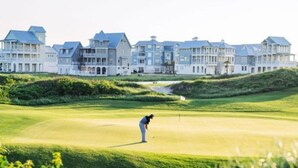 A golf course with a person playing, surrounded by a large, modern building with multiple balconies. - Townhouse + loft with private balcony, heated pool, gym, beach access & W/D (Port Aransas)