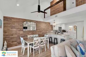 A modern kitchen with white cabinets and a central island, a dining area with white chairs, and a ceiling fan.