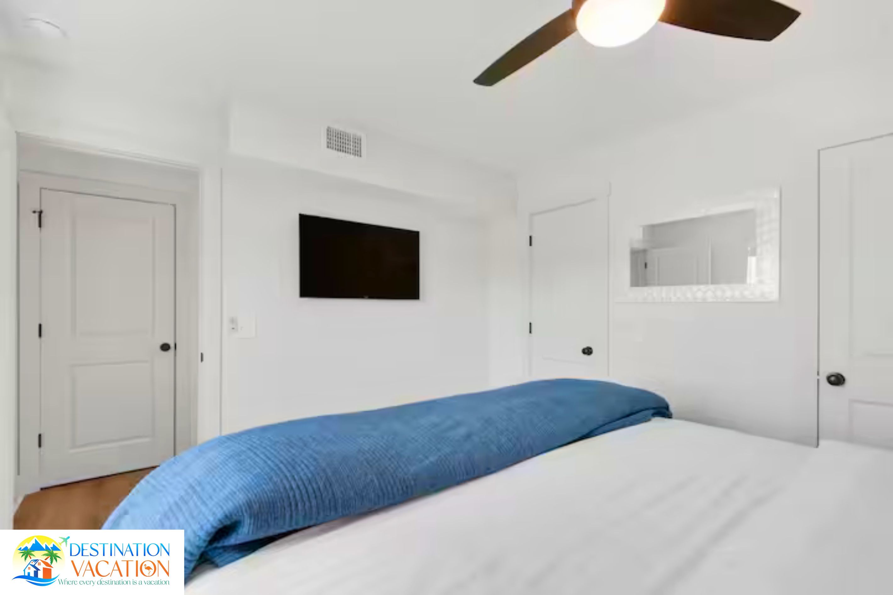 A hotel room with a bed, a ceiling fan, a television, and a mirror.