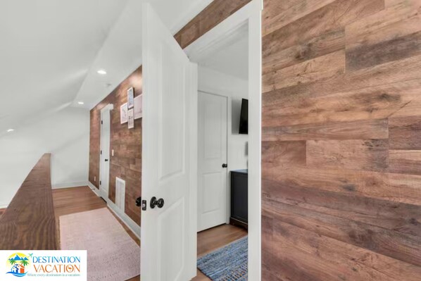 Un passadís amb terres de fusta, parets blanques i una porta que dona a una habitació amb televisió.