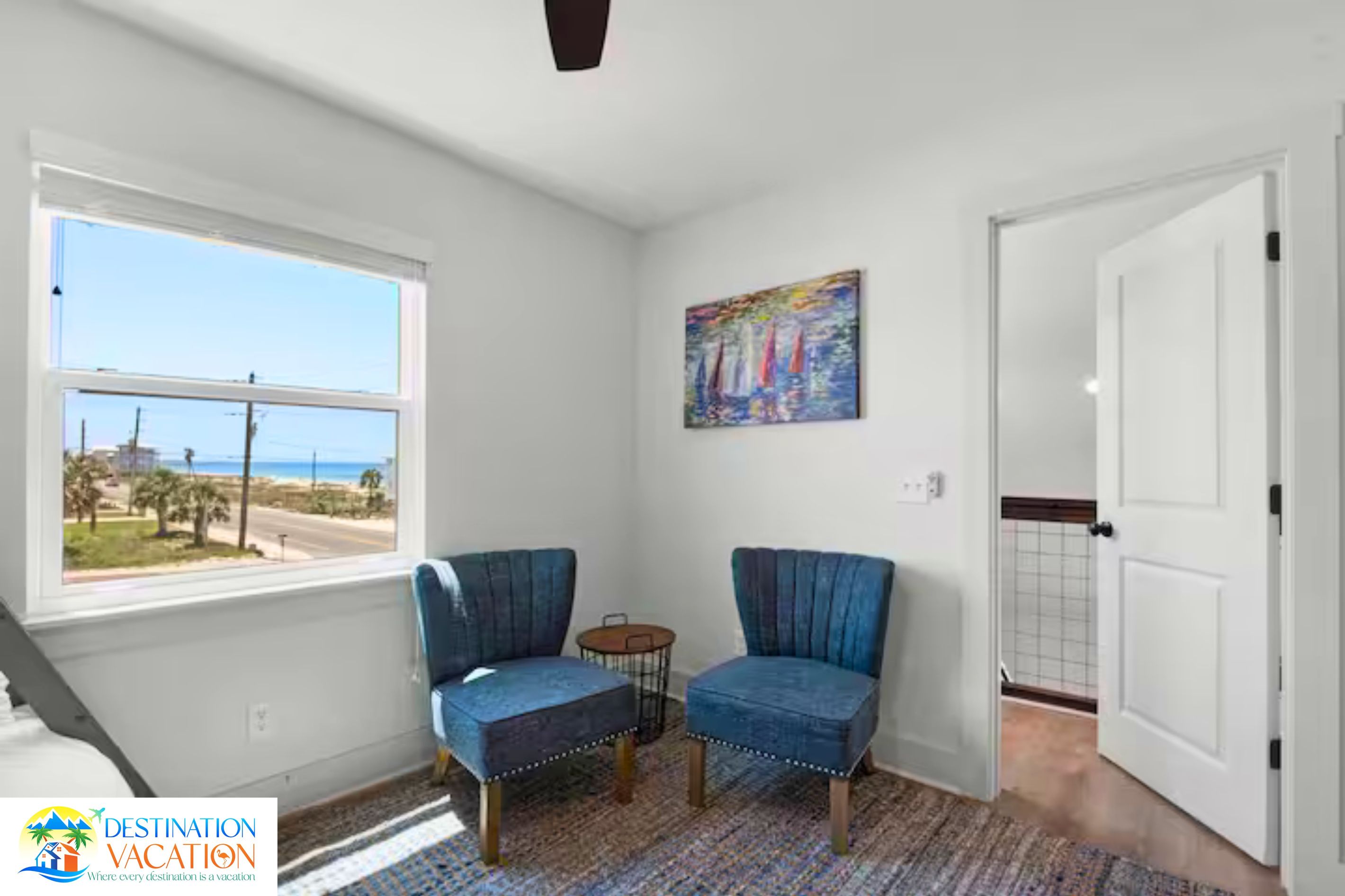A room with two blue chairs, a small table, a painting on the wall, and a view of the beach through a window.