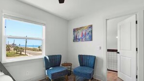 Una habitació amb dues butaques blaves, una taula rodona petita, un quadre a la paret i una finestra amb vista a la platja.