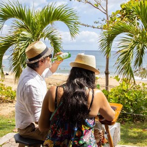 Deux personnes sont assises sur un banc, face à l’océan, avec des palmiers et une plage en arrière-plan.