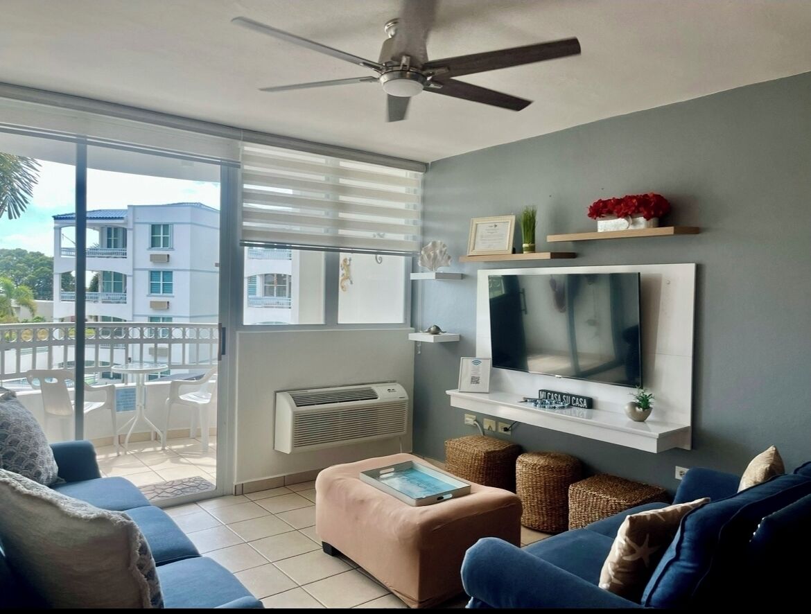 Un salón con televisor, sofá, reposapiés y un balcón con vistas a edificios.