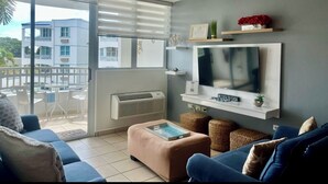 A living room with a TV, sofa, ottoman, and a balcony with a view of buildings. - Gated condo, pool, high speed Wi-fi, washer & dryer. Walk to beach, near town  (Rincon)