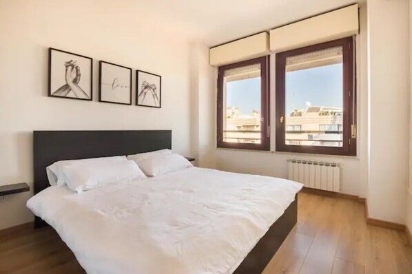 Une chambre avec un grand lit, une fenêtre donnant sur des immeubles et trois tableaux encadrés accrochés au mur.