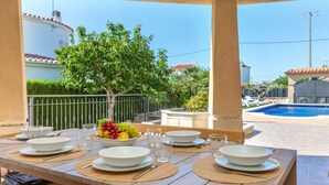 An outdoor dining area with a table set for four, a pool in the background, and a tree providing shade.