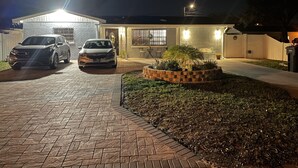 Et hus med to biler parkeret i indkørslen, en veloplyst forhave og en brostensbelagt sti, der fører til indgangen.