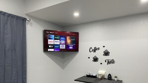 Et moderne værelse med et fladskærms-tv på væggen, der viser forskellige streamingtjenester. Der er et lille bord med barstole, og en klistermærke på væggen med teksten "Coffee Time".